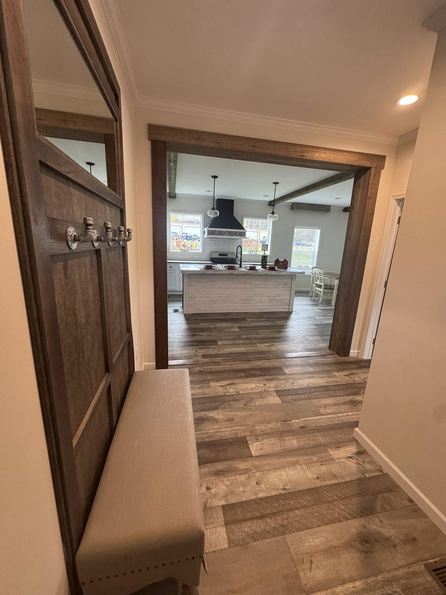 Hallway leading to a cozy kitchen with wooden floors and rustic decor. The kitchen features a central island, pendant lights, and a large hood.