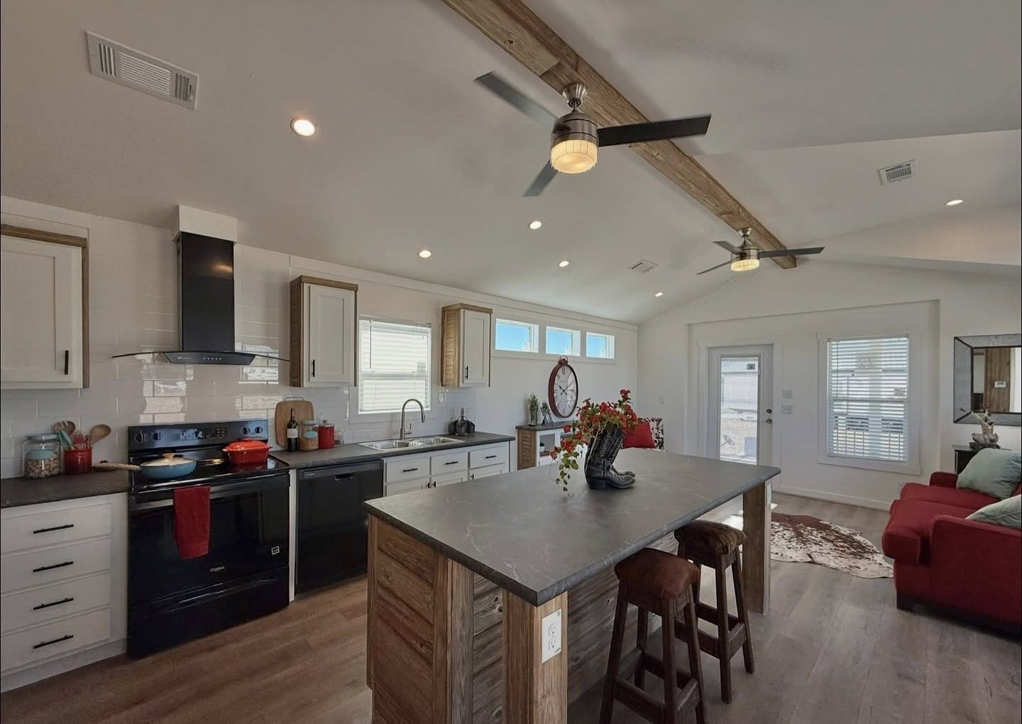 Spacious kitchen-living area with rustic wood beams, modern appliances, center island, bar stools, red sofa, and natural light. Cozy and inviting atmosphere.