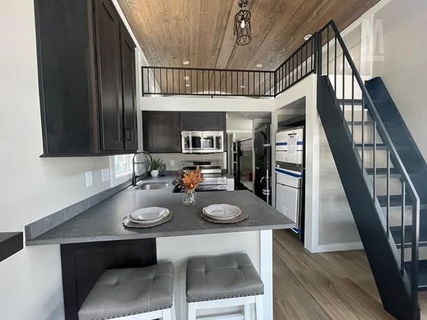 Modern kitchen with dark cabinets, gray countertops, and a high ceiling with wood paneling. A staircase leads to a loft. Cozy, organized space.