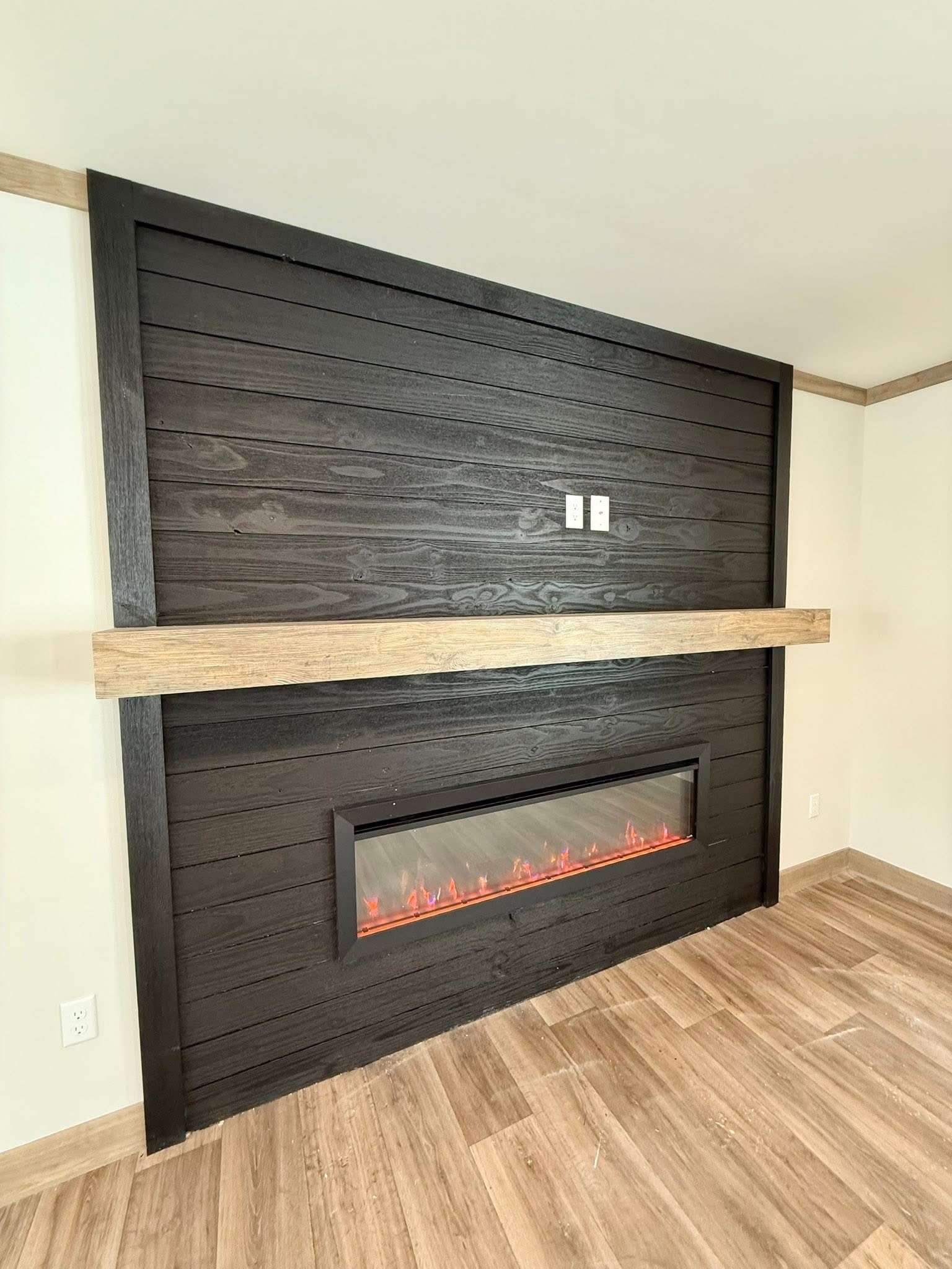 Modern fireplace with glowing embers set in a dark wooden shiplap wall, accented by a light wood mantel. The room has light wooden flooring.