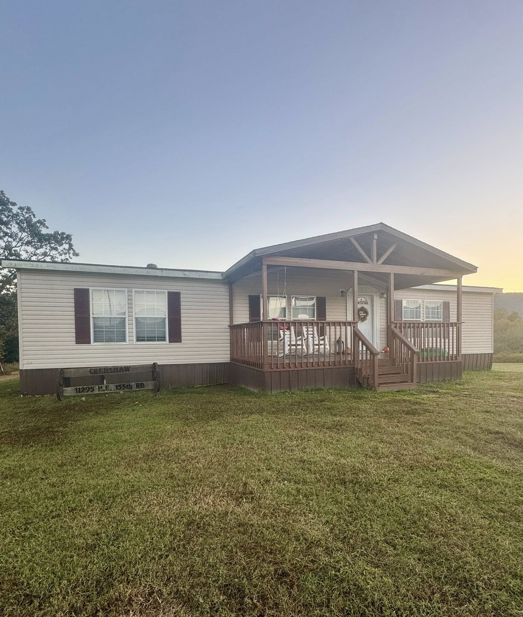 Single-story house with a front porch and brown shutters, set against a clear evening sky. A wide lawn surrounds the home, evoking a serene rural ambiance.