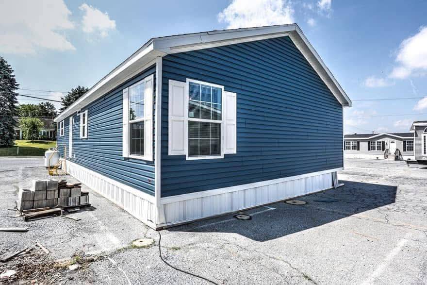 Blue mobile home with white trim and windows, situated on a paved lot under a clear sky. Nearby are stacked concrete blocks and other mobile homes.