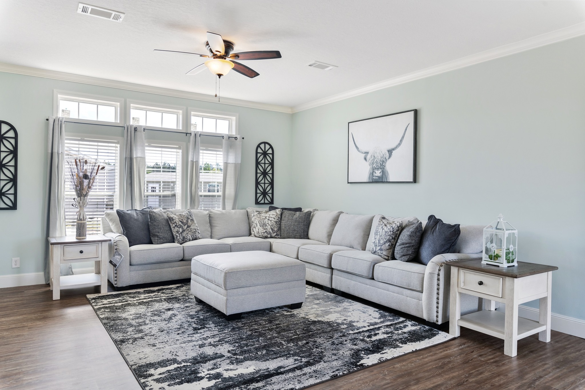Spacious living room with light gray sectional couch, abstract rug, and art of a bull. Soft blue walls and large windows create a bright, serene atmosphere.