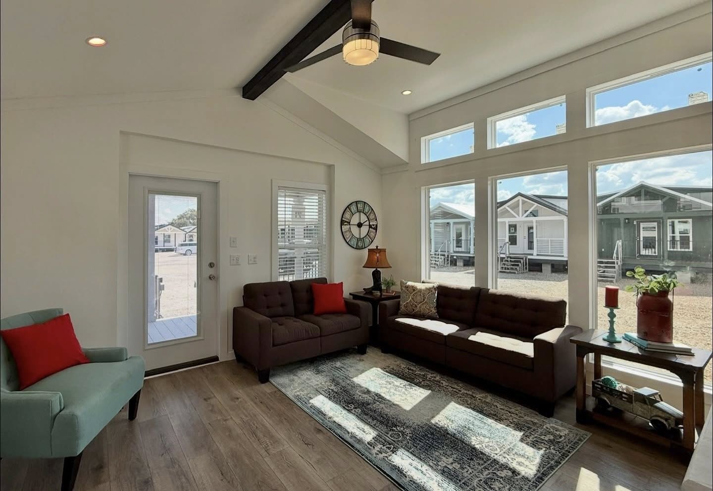 Bright living room with modern decor, featuring a teal chair, brown sofas with red pillows, and large windows showing tiny houses outside. Cozy and inviting.