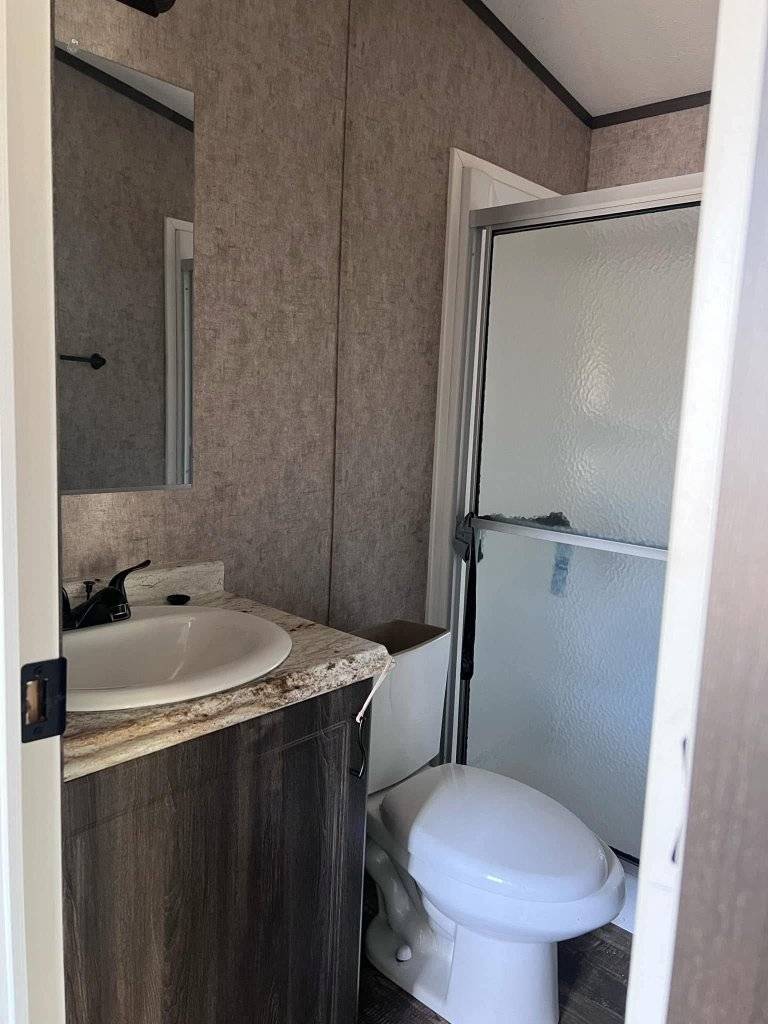Small bathroom with earth-toned wallpaper featuring a white toilet, a wood vanity with a marble countertop, a sink, and a glass shower door.