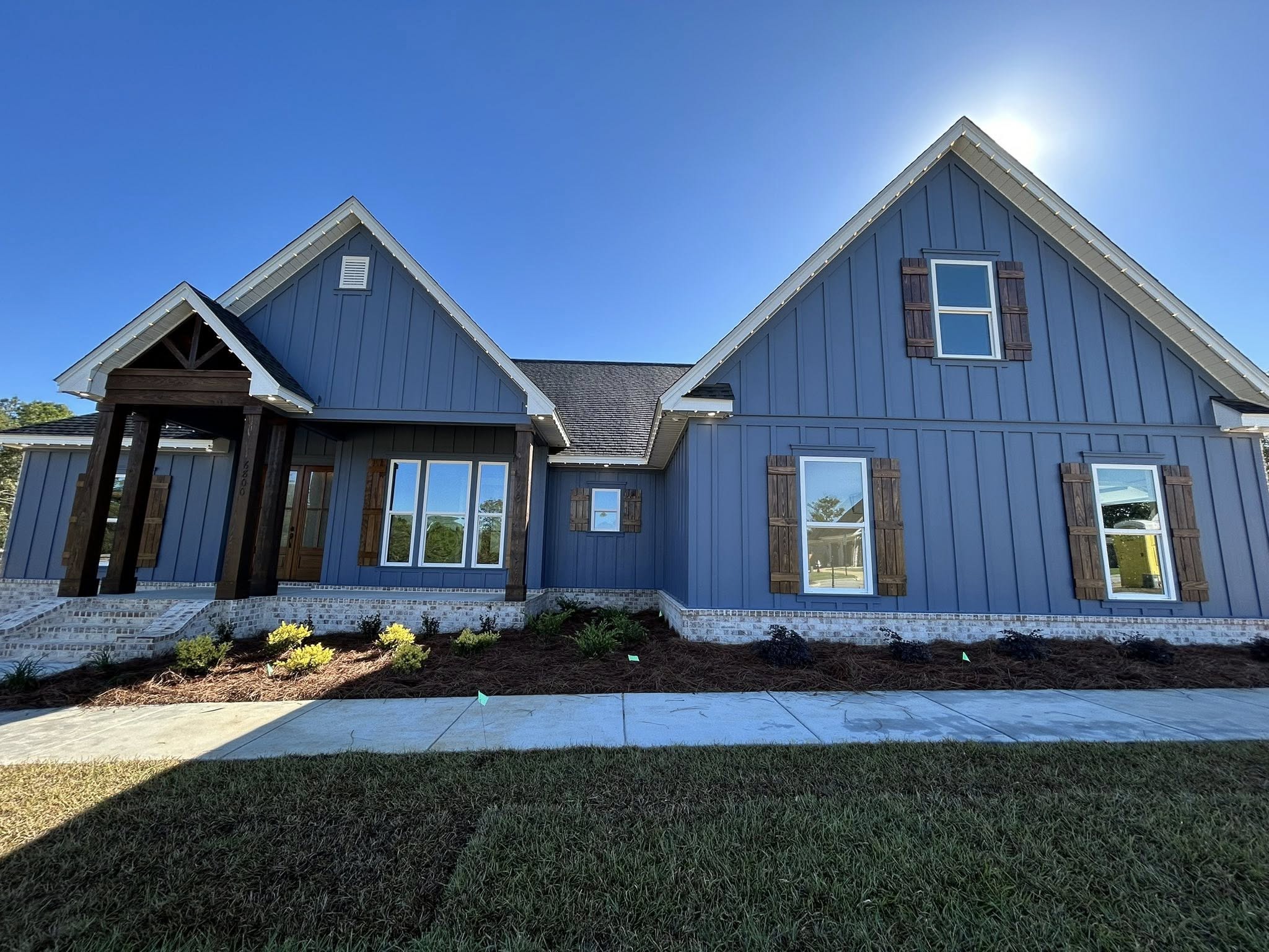 A modern blue house with wooden shutters and a pitched roof. Manicured lawn and shrubs in front, under a clear blue sky with bright sunlight.