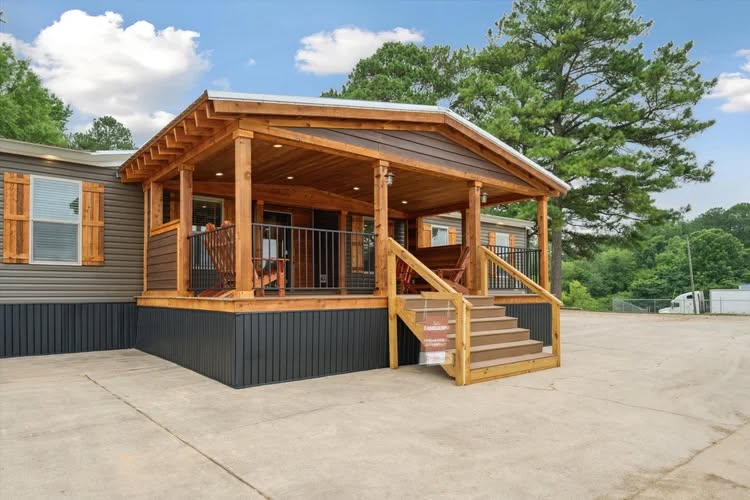 A cozy wooden porch attached to a modern house with a sloped roof, surrounded by lush greenery and a clear blue sky, conveying tranquility.
