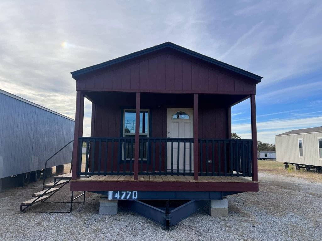 Compact tiny house with a red exterior, porch with railing, and white door, set on a gravel lot under a partly cloudy sky, conveying simplicity and neatness.