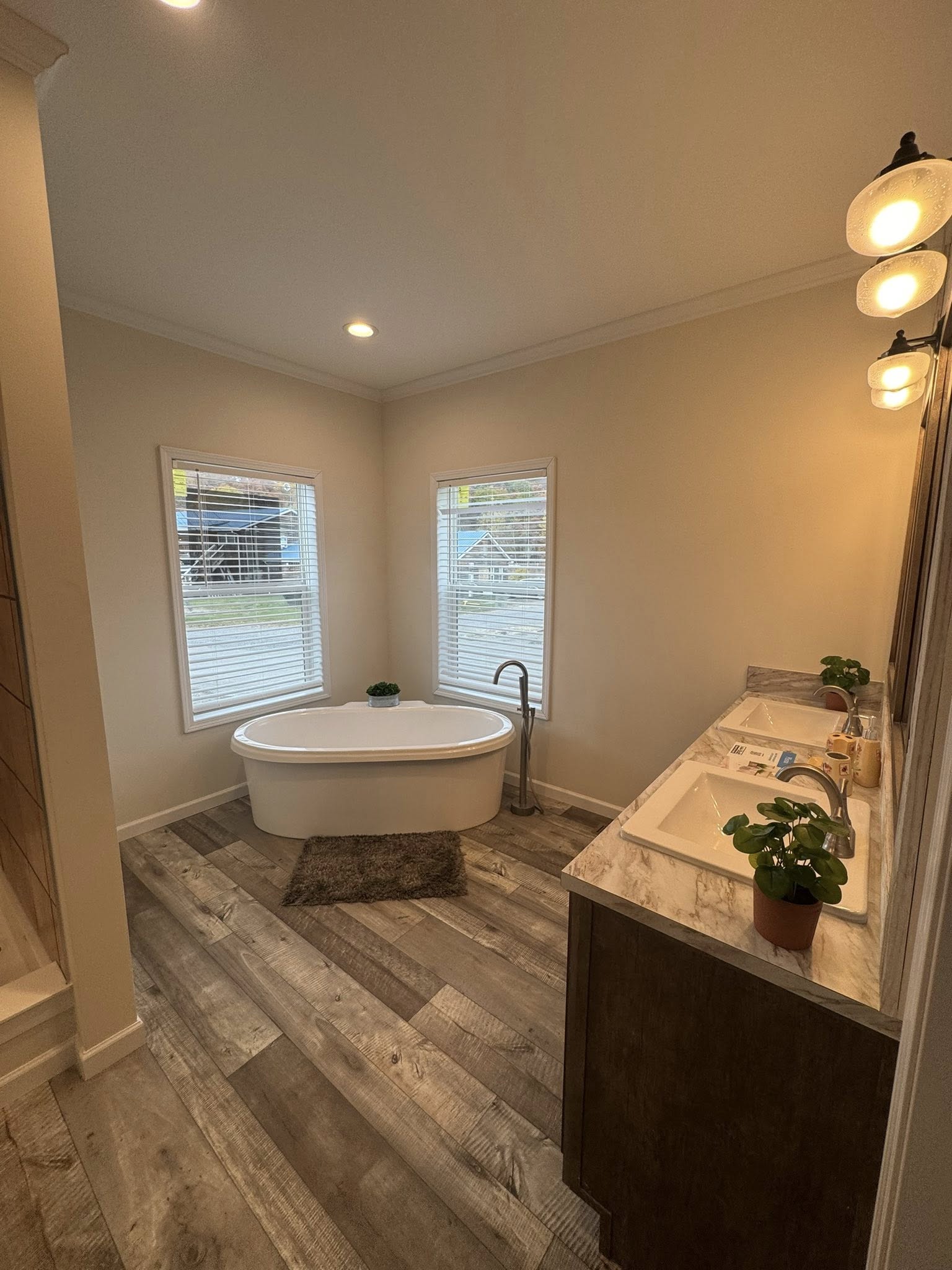 Stylish bathroom with a freestanding oval tub by two windows. Wooden floorboards, marble-topped vanity, and soft lighting create a serene atmosphere.