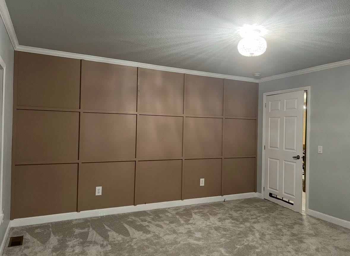 A room with a light brown paneled accent wall, gray carpeting, and a white door on the right. A bright ceiling light casts a soft, warm glow.