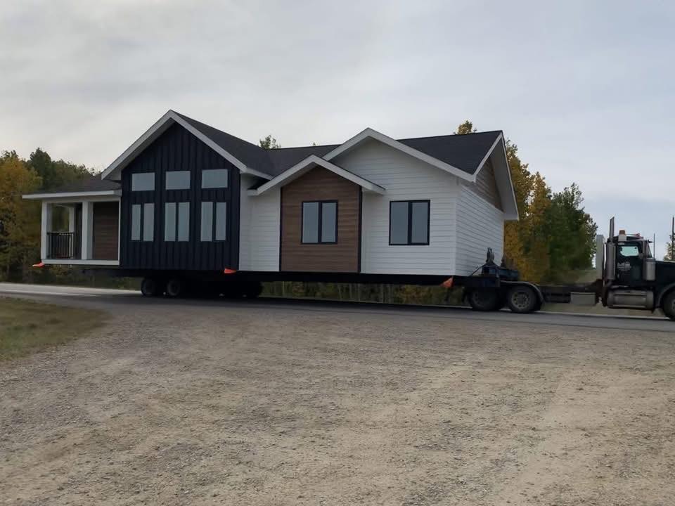 A large house, painted white with dark panels, is transported on a flatbed truck along a rural road. The scene is calm with trees in the background.