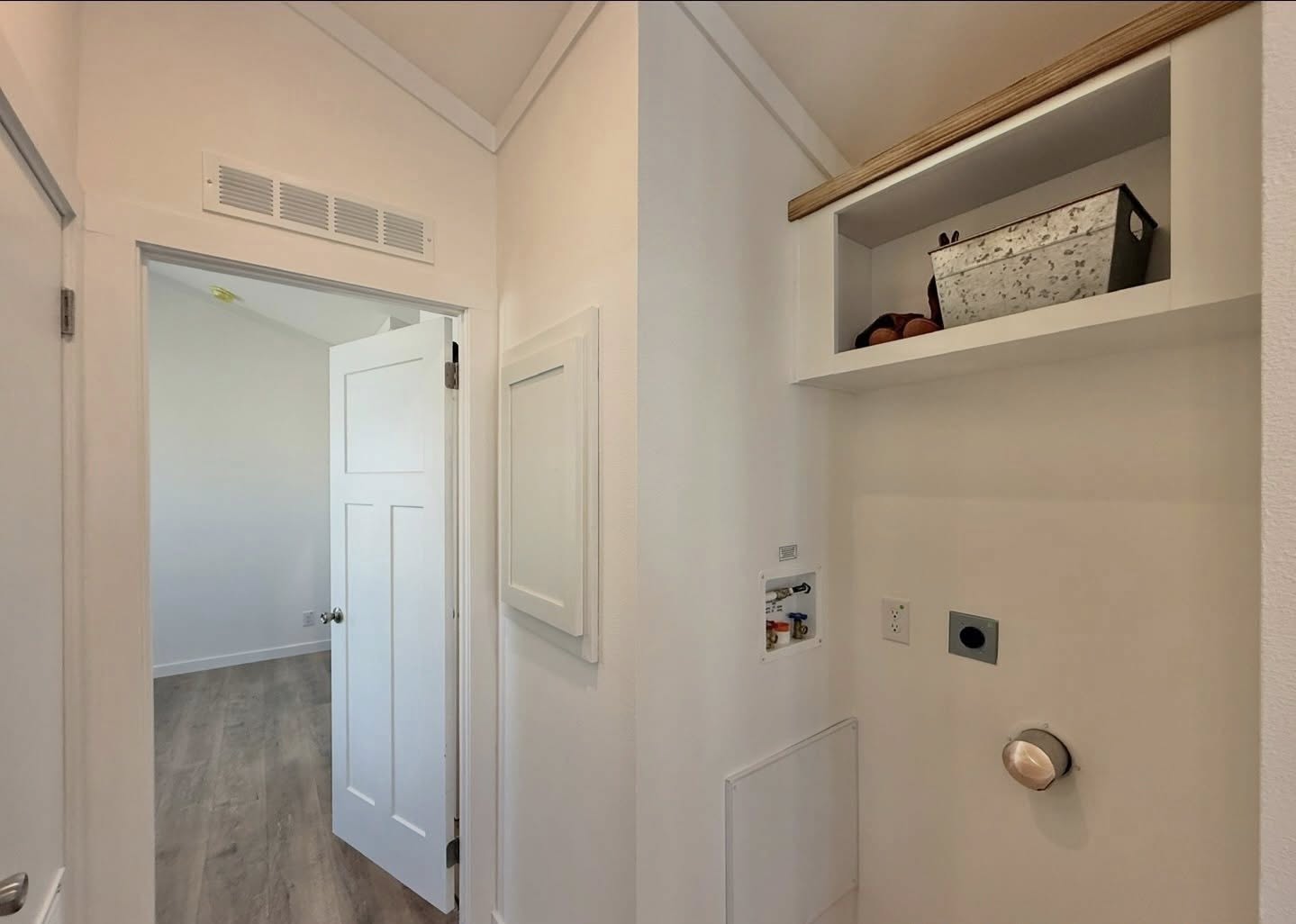 Bright hallway with light wood flooring, open white door, and built-in shelf holding a metal box. Cozy and minimalistic ambiance.