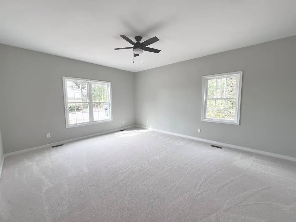 Empty room with light gray walls and white carpet. Two windows allow natural light in. A ceiling fan hangs from the center, creating a calm atmosphere.