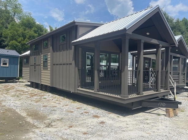 A modern tiny house with a metal roof and a covered porch, situated on a gravel lot. Large windows and a neutral color palette create a cozy feel.