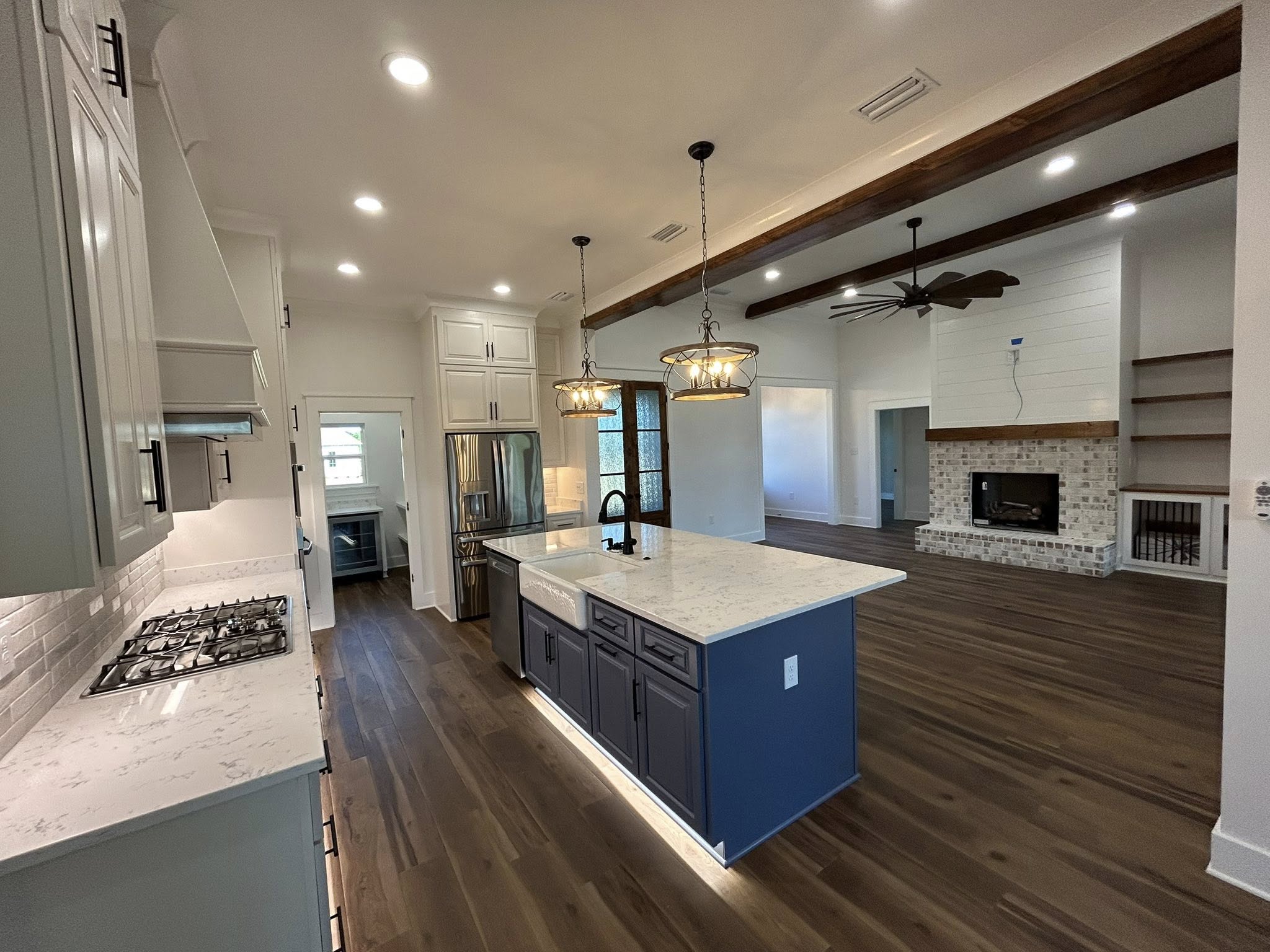 Spacious modern kitchen and living area with hardwood floors, white cabinets, marble countertops, and a central island. Two pendant lights hang above the island; a cozy fireplace sits in the living space, exuding warmth.