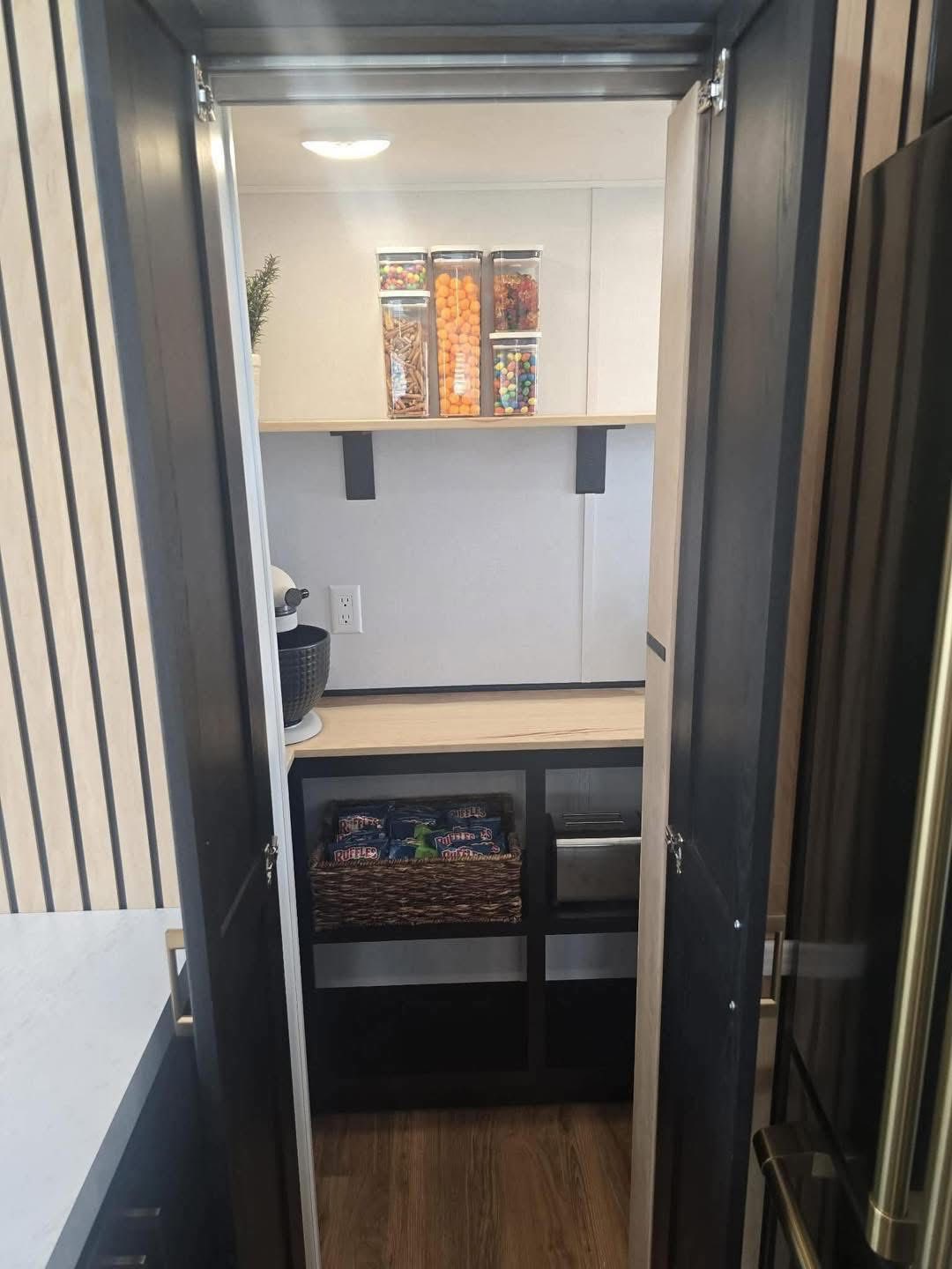 Compact pantry with open double doors, featuring organized shelves with jars of snacks and candies. A small appliance sits on the left counter.