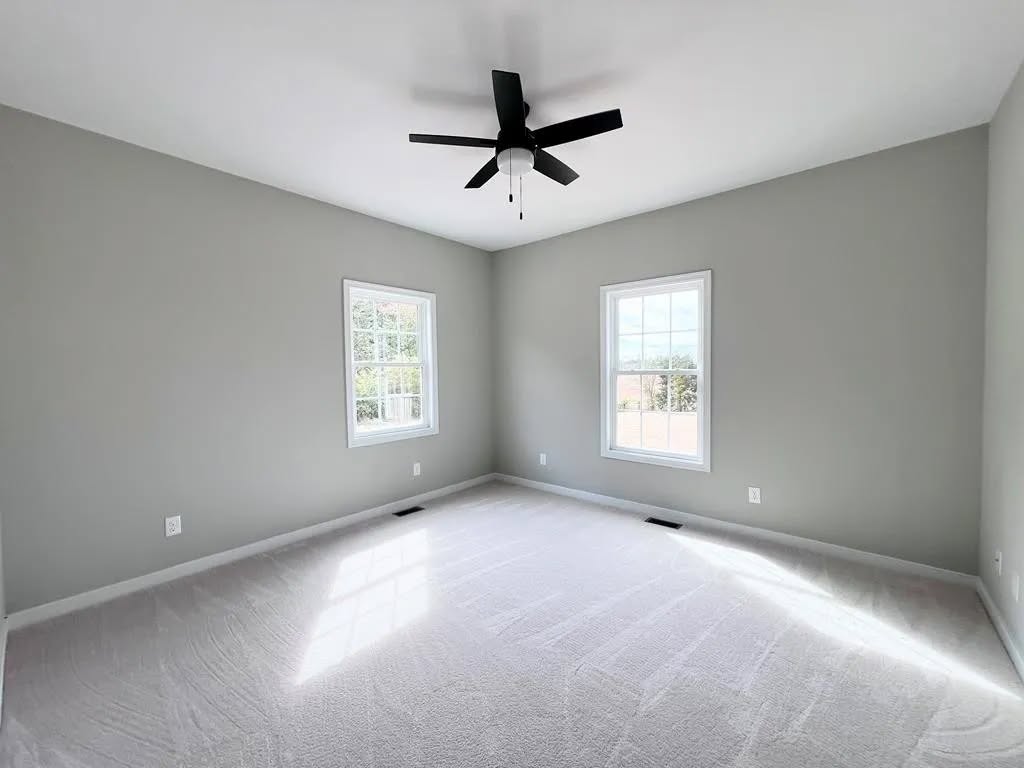 A bright, empty room with light gray walls, white trim, and a white carpet. Two windows provide natural light. A black ceiling fan is centered on the ceiling.