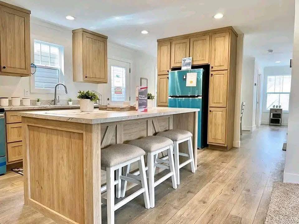 Modern kitchen with light wood cabinets, a turquoise fridge, and a central island featuring stools and plants. Bright, airy, and welcoming atmosphere.