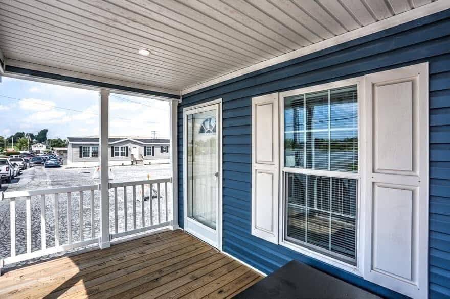 Covered porch with wooden floor, blue siding, and white trim. Glass door and window reveal a sunny, inviting ambiance. Parking lot visible outside.