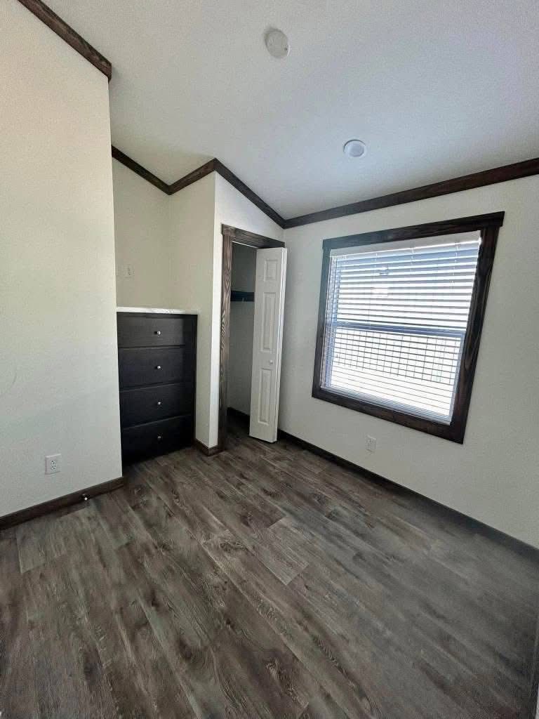 Small, empty room with wooden floors, a large window with blinds, and a built-in dark dresser. An open closet door adds depth and light.