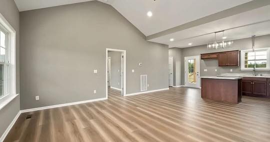 Spacious, modern open-plan room with light wood flooring and gray walls. Features a kitchen with dark cabinets and island, under bright ceiling lights.