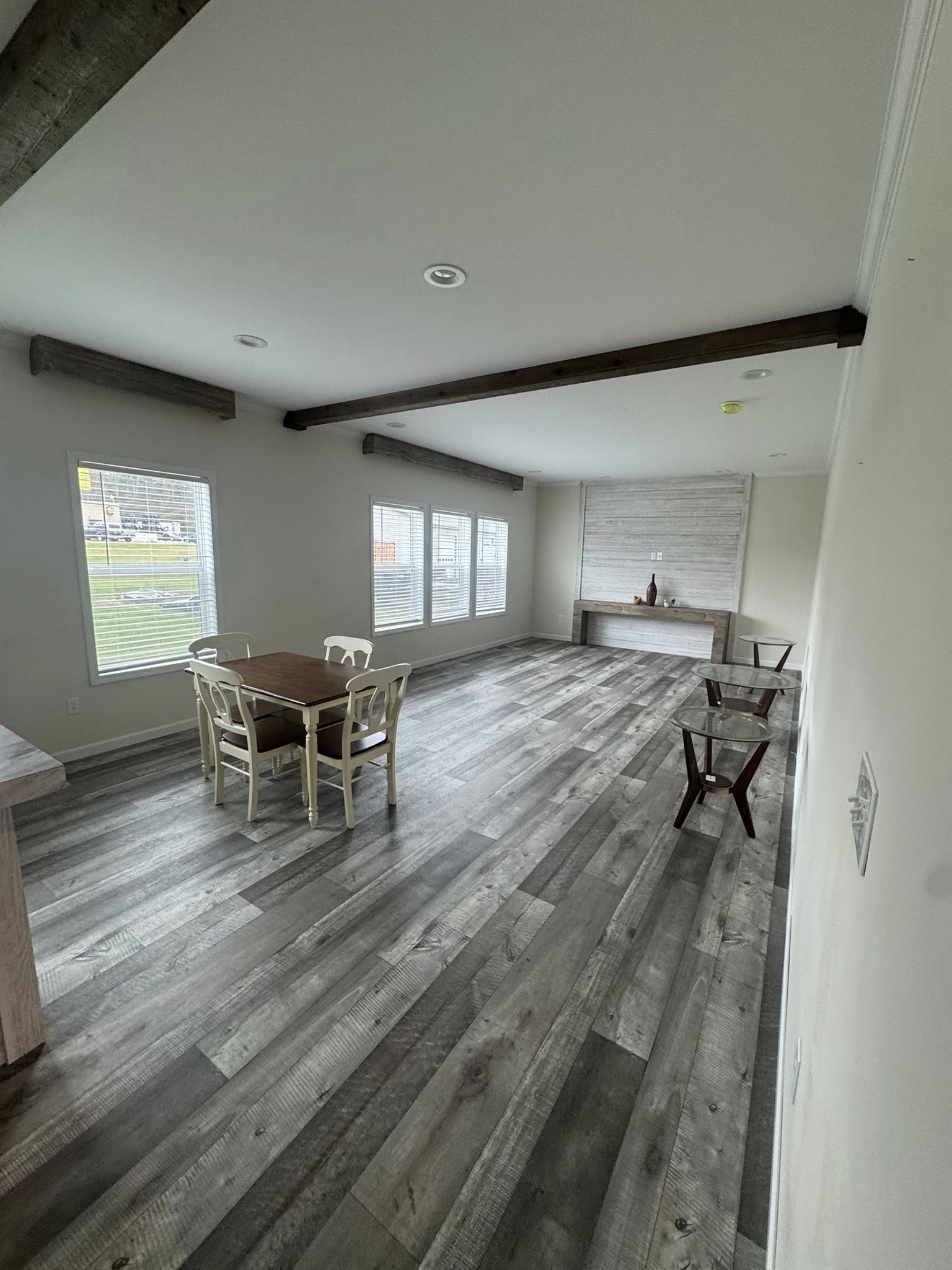 Spacious modern room with wood floors and ceiling beams. A small dining table with four chairs is near two large windows, creating a bright ambiance.