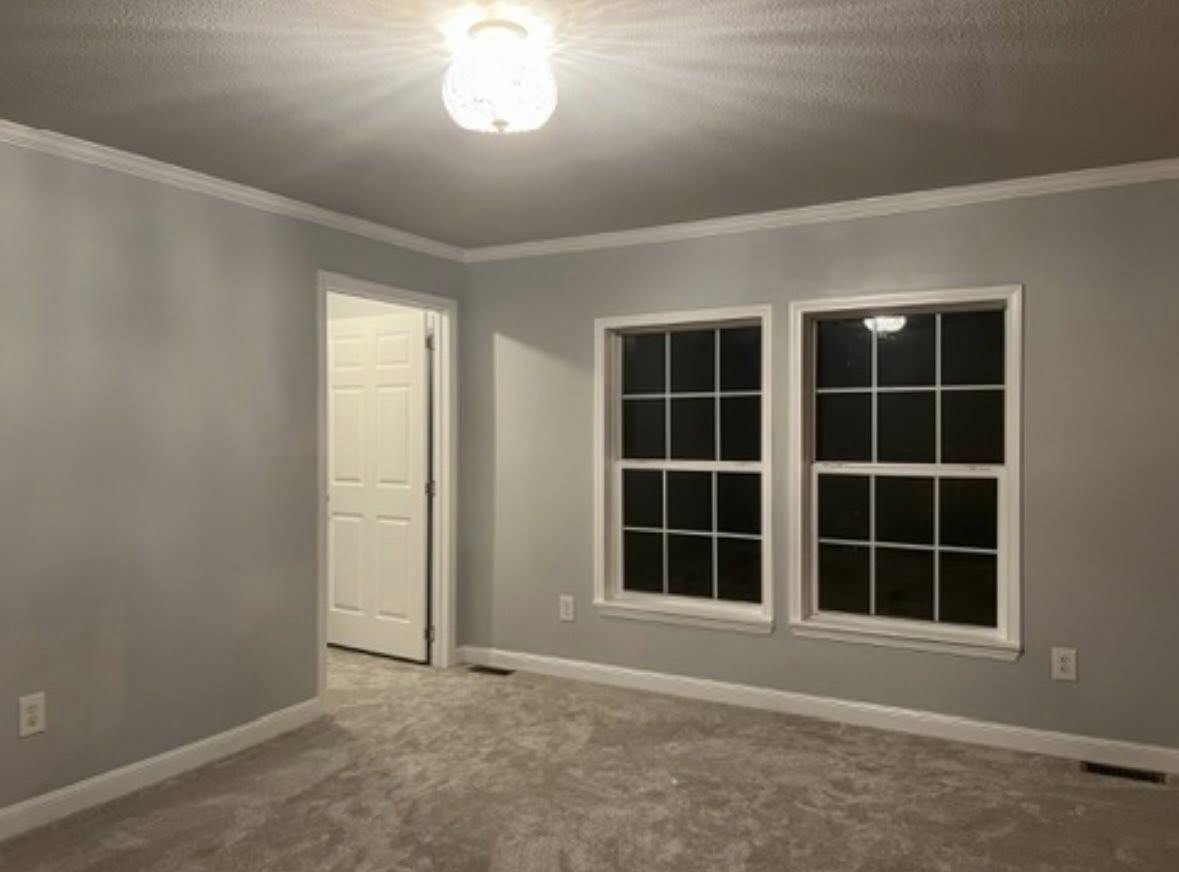A dimly lit, empty room with gray walls and carpet. Features two large windows, a simple ceiling light, and an open doorway to the left, conveying a calm atmosphere.