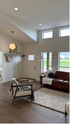 Bright living room with high ceiling, featuring a brown leather sofa, modern chair with a blanket, large windows, and a beige rug. Cozy and inviting atmosphere.
