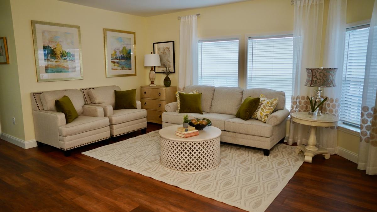 A cozy living room features a beige sofa and chairs with green cushions, a round patterned coffee table on a light rug, framed art, and wooden floors.