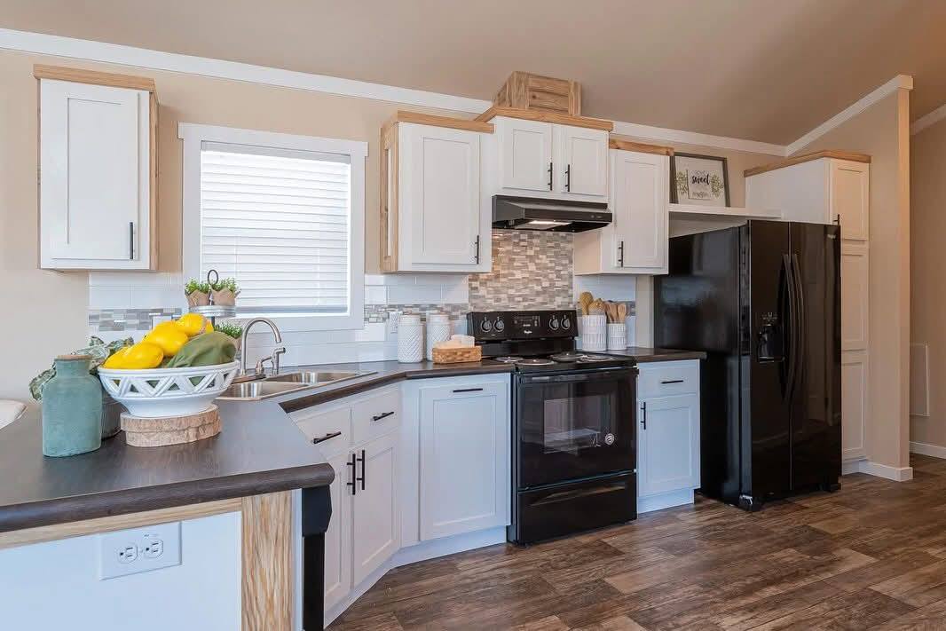 Bright kitchen with white cabinets and black appliances. A bowl of fruits adds color, while wood accents and a backsplash create warmth. Modern and inviting.