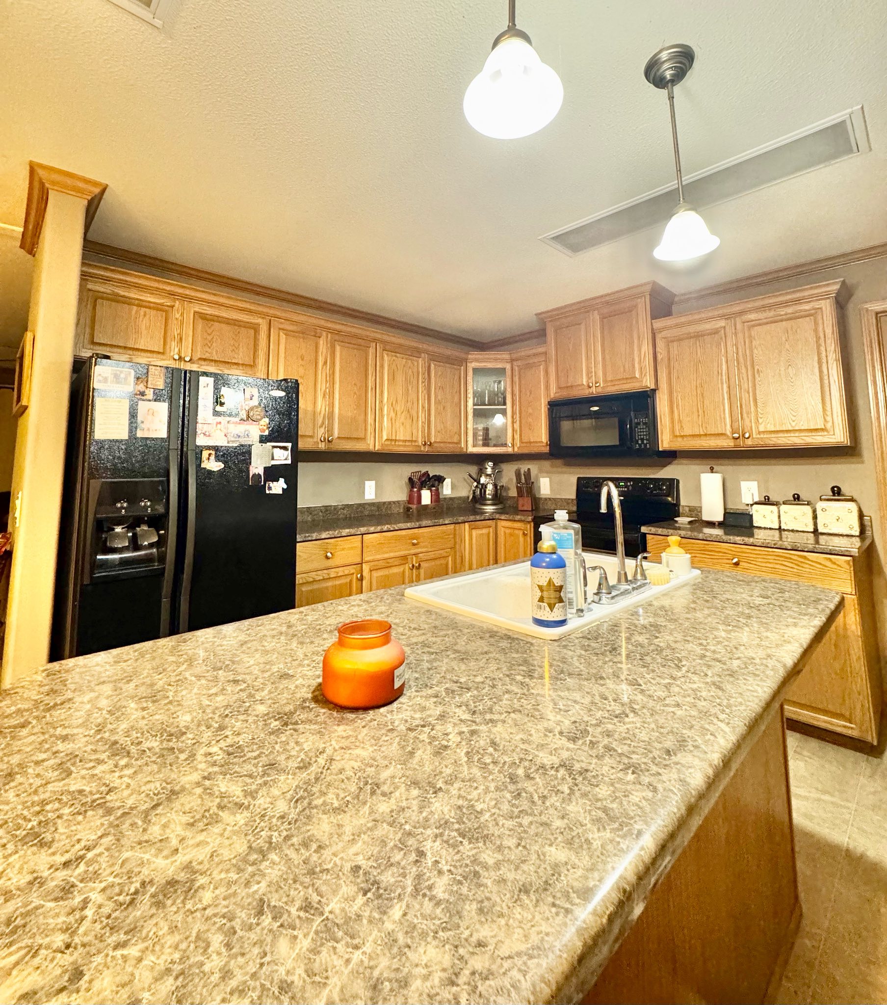 Warm kitchen with wooden cabinets, granite countertops, and black appliances. Orange candle on island; cozy and inviting atmosphere.