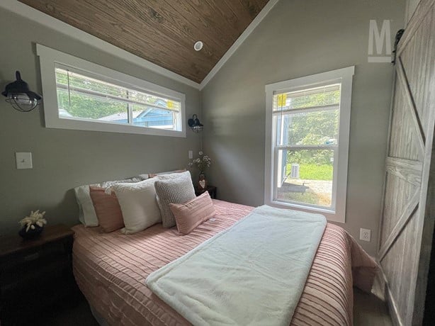 Cozy bedroom with sloped ceiling, featuring a bed with pink bedding and multiple pillows. Two windows let in natural light, creating a serene atmosphere.