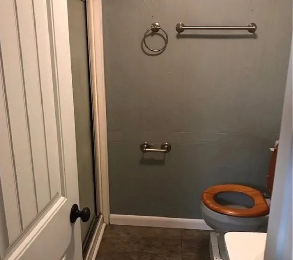 Small bathroom with gray walls, white door, and a wooden toilet seat beside a glass shower door. Towel racks on wall; cozy yet cramped space.