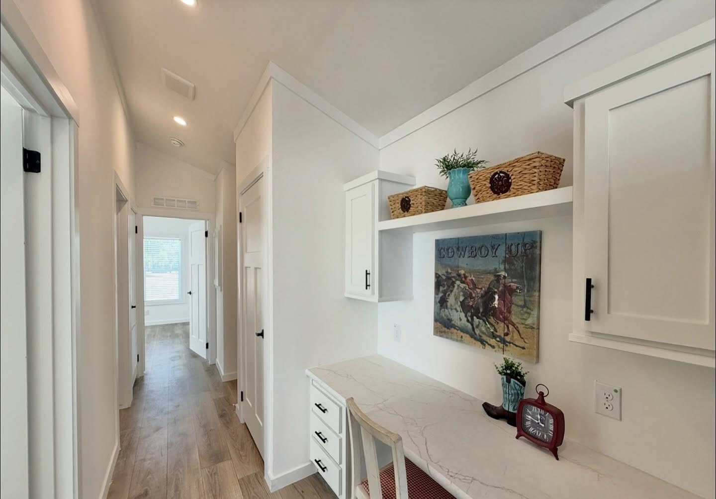 Bright hallway with light wooden flooring, white walls, and a built-in desk with cabinets. Decor includes a cowboy painting, red clock, and wicker baskets.