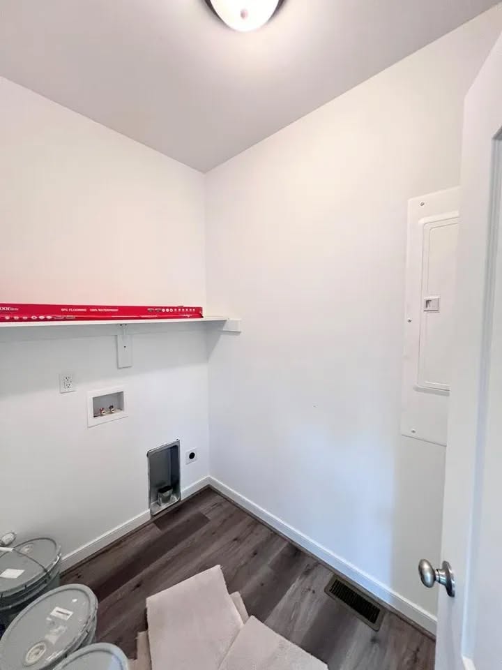 Small, empty laundry room with white walls and a wood floor. A shelf holds paint cans and a level. Plumbing fixtures are visible, and light from a ceiling fixture brightens the space.
