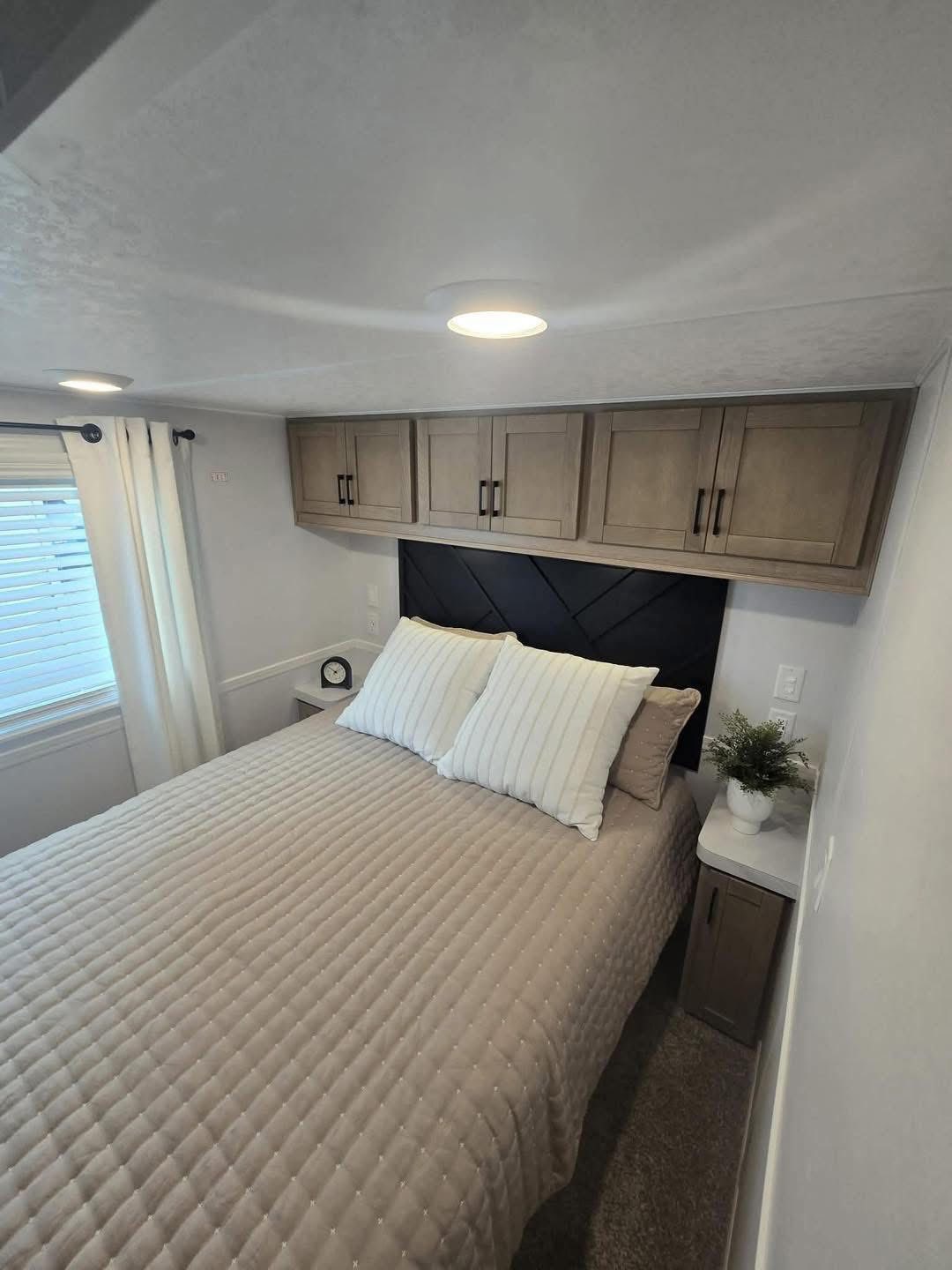 A cozy bedroom featuring a neatly made bed with a beige quilt and two striped pillows. Wooden cabinets and a small plant provide a warm, inviting atmosphere.