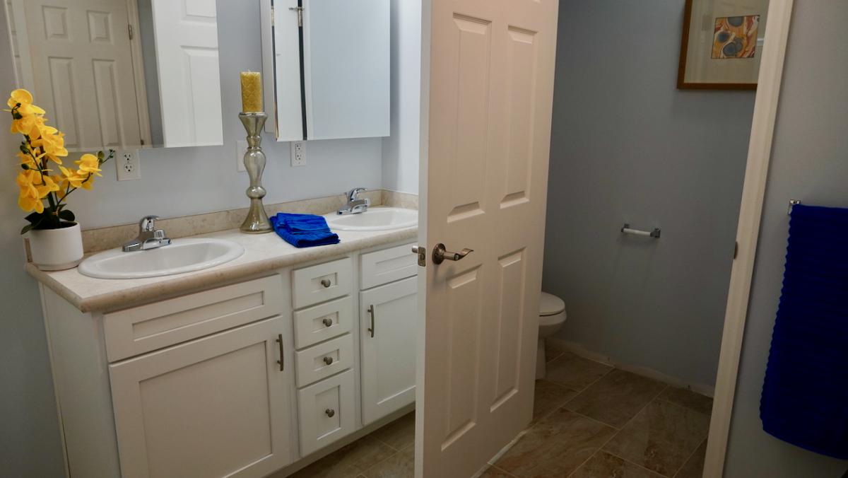 A clean, modern bathroom features a double sink vanity with white cabinets, a yellow orchid, and blue towels. An open door reveals a toilet.