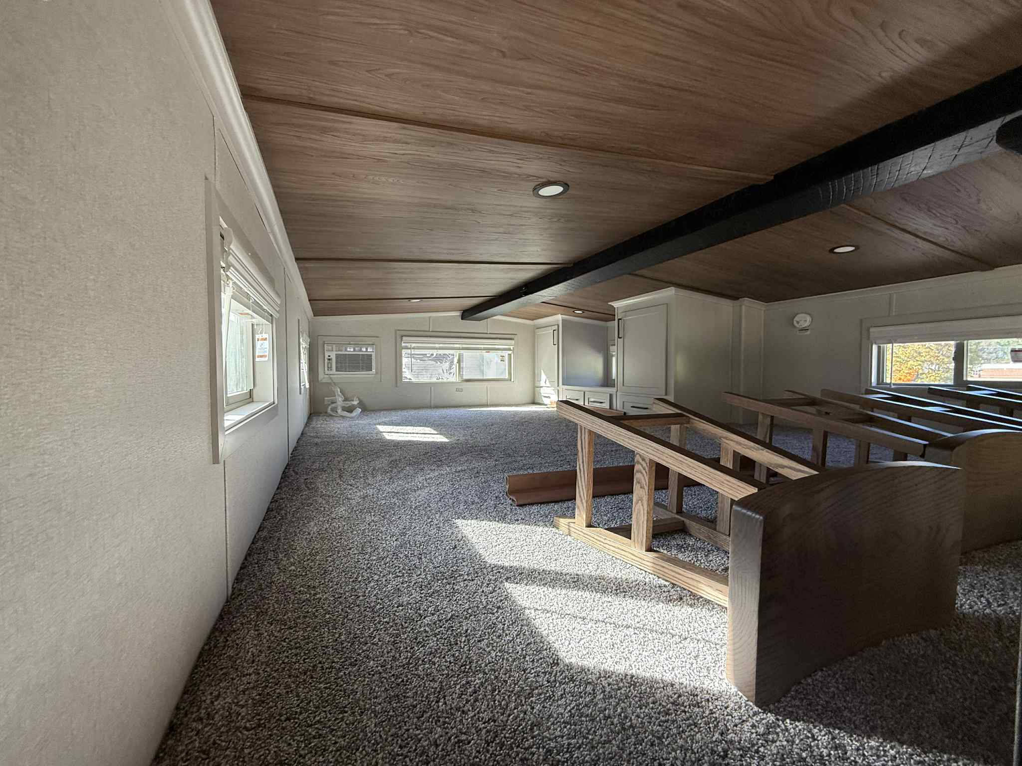 Cozy attic room with carpeted floor and wooden ceiling, featuring benches along the side. Sunlight streams through windows, creating a warm ambiance.