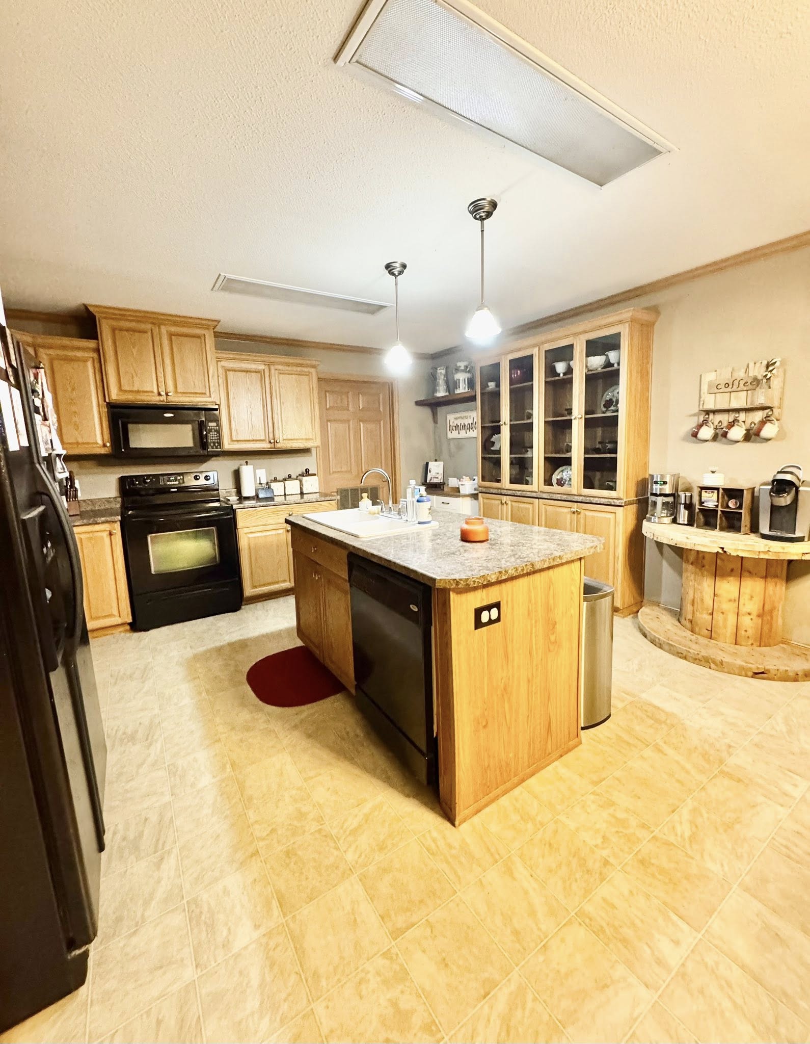 A cozy kitchen with wooden cabinets, an island with a granite countertop, and pendant lights. Black appliances and a rustic coffee station create a warm, inviting atmosphere.