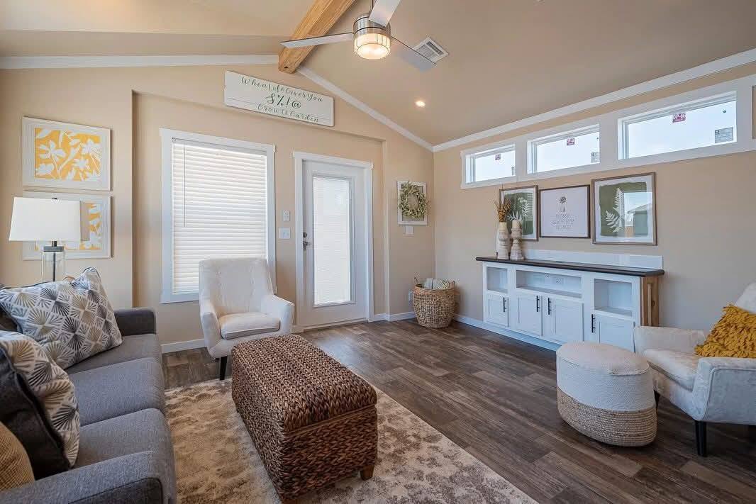 Cozy living room with warm tones, featuring a gray sofa, wicker ottoman, and armchairs. Wooden floors, wall art, ceiling beams, and a white cabinet complete the space.
