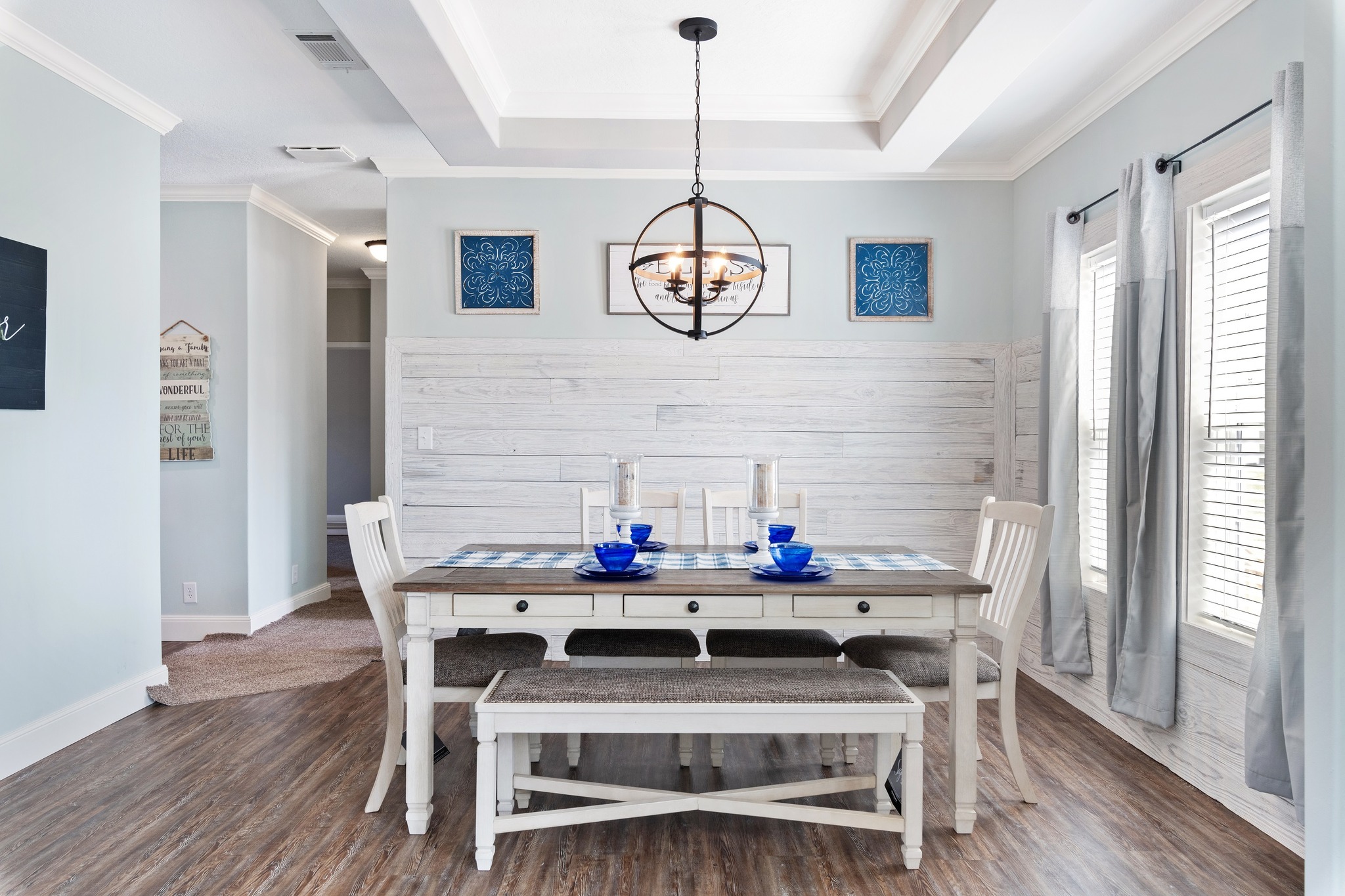 Bright dining room with a rustic wooden table, white chairs, and blue accents. A modern chandelier hangs overhead. Sunlight streams through large windows.