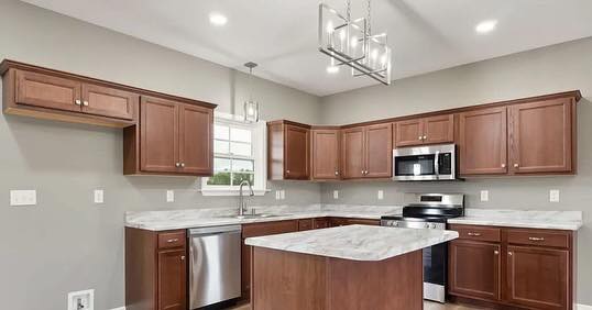 Modern kitchen with wooden cabinets, stainless steel appliances, and a marble-patterned island. Pendant light and a window above the sink add brightness.