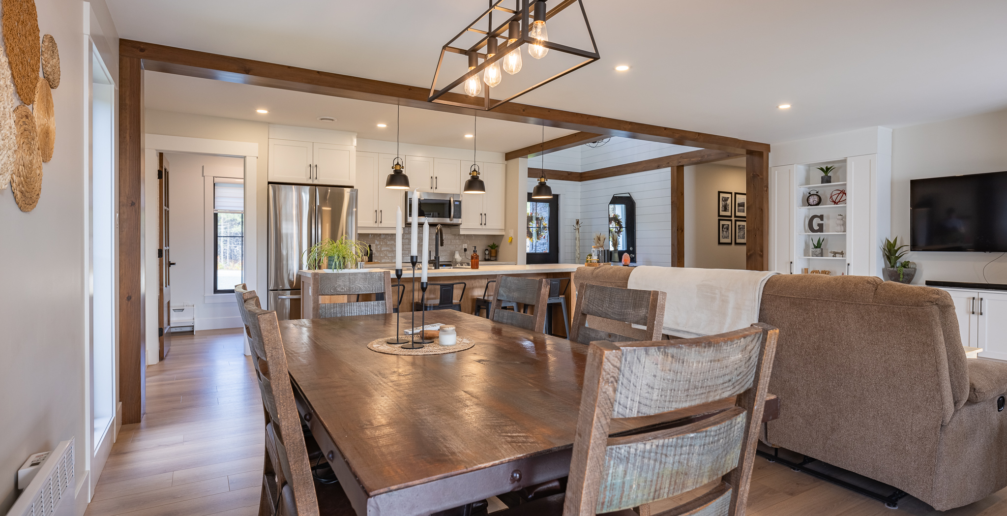 Spacious open-plan living area with a rustic wooden dining table, modern kitchen, brown couch, and minimalist decor. Bright, inviting ambiance.