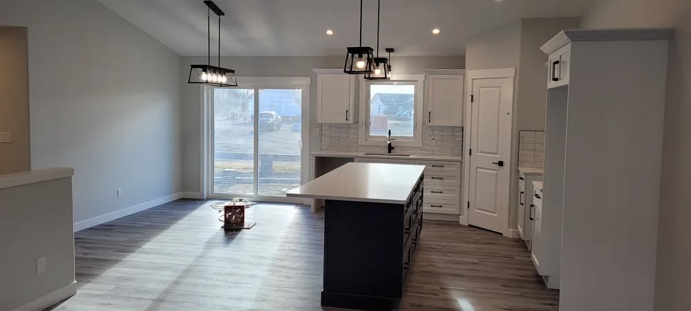 Spacious kitchen with an island, white cabinets, and wood flooring. Sunlight streams through large sliding doors, highlighting pendant lights.