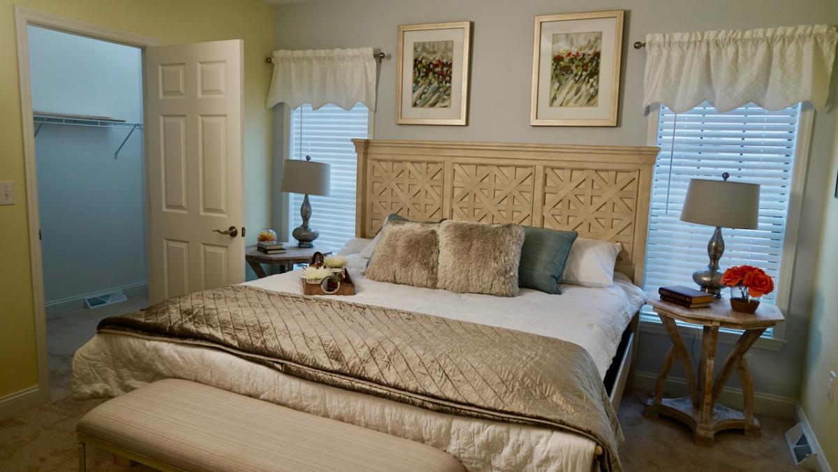 Cozy bedroom with a cream and beige palette, featuring a patterned headboard, plush pillows, two bedside tables with lamps, and floral artwork above.