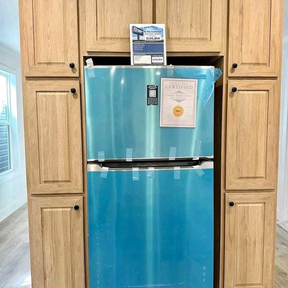 A refrigerator wrapped in blue protective film is incompletely installed in a wood cabinet. Price tag and certification are affixed on the door.