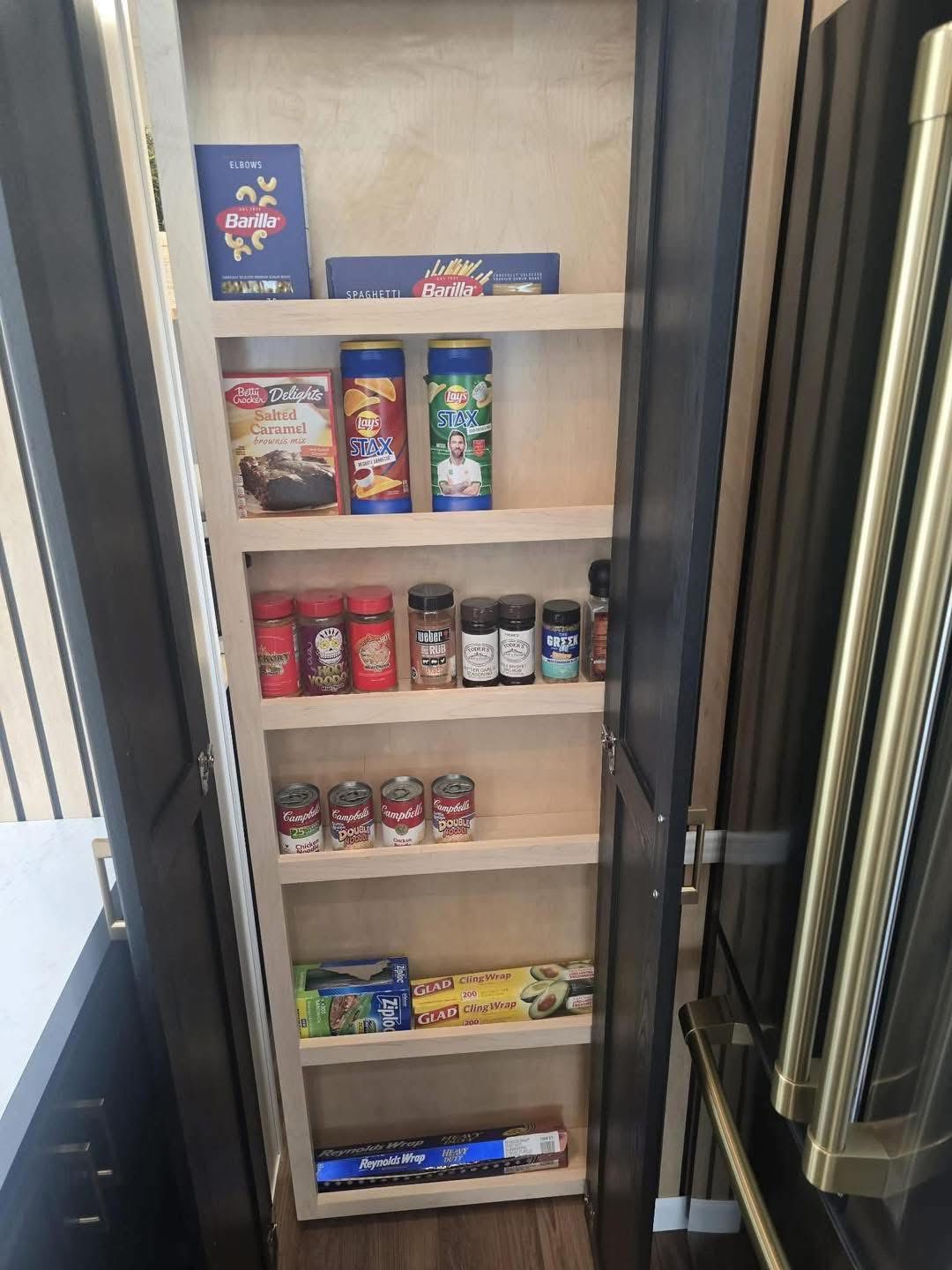 Tall pantry cabinet with five shelves holding pasta boxes, canned goods, chips, spices, and wraps. Modern kitchen setting with an organized, tidy feel.