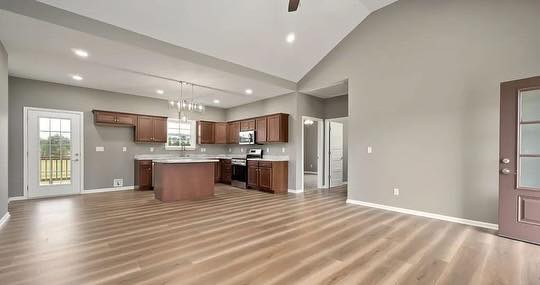Spacious open-plan room with light wood flooring, gray walls, and high ceiling. It features a kitchen with brown cabinets and an island under modern lighting.