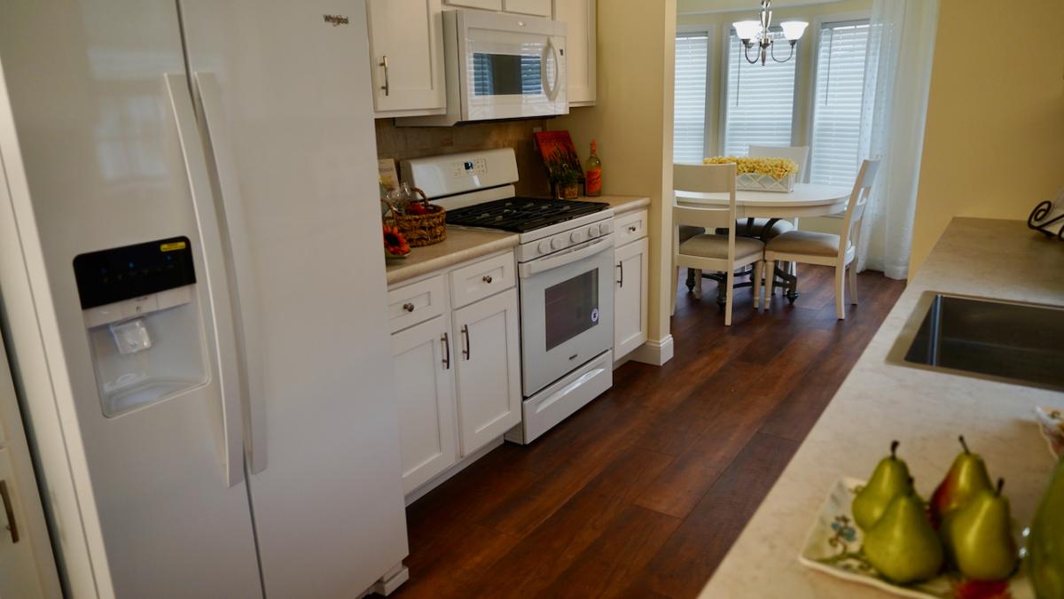Bright kitchen with white cabinets, a fridge, and a stove. A fruit basket and pears decorate the counters. The dining area is in the background, featuring a round table and a chandelier.
