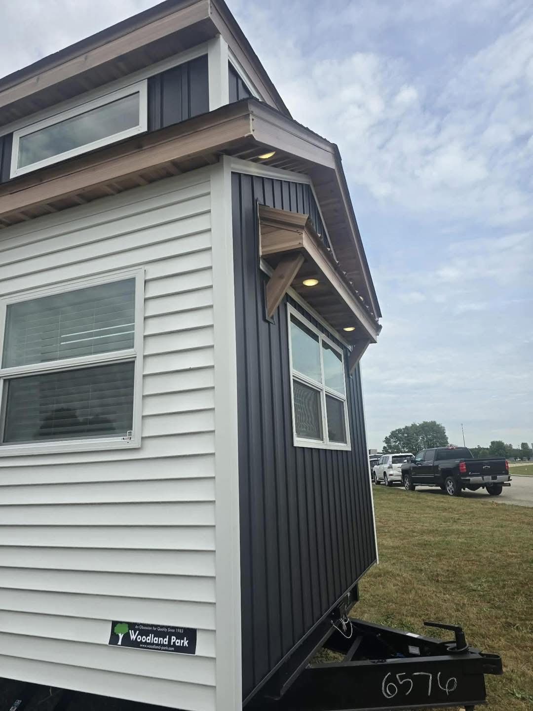 A small mobile home with black and white siding stands on a grassy area. It has a wood-accented roof and windows. The sky is overcast, conveying a calm mood.
